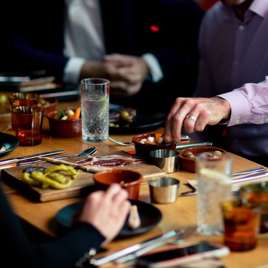 A group of people seen from the shoulder down seated at a dining table with an assortment of small Spanish sharing plates, each taking pieces from different dishes.